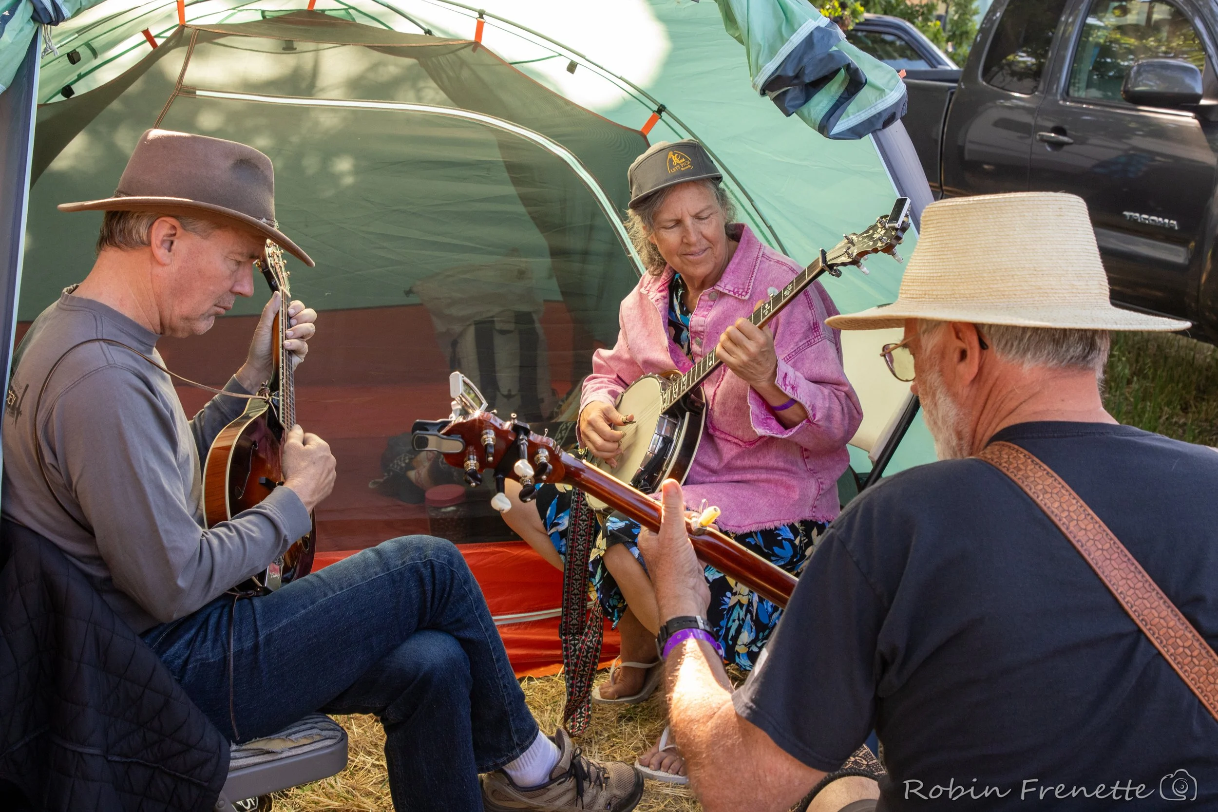 Three adults sitting outside under a green camping tent, playing musical instruments together at the Parkfield Bluegrass Festival campsite