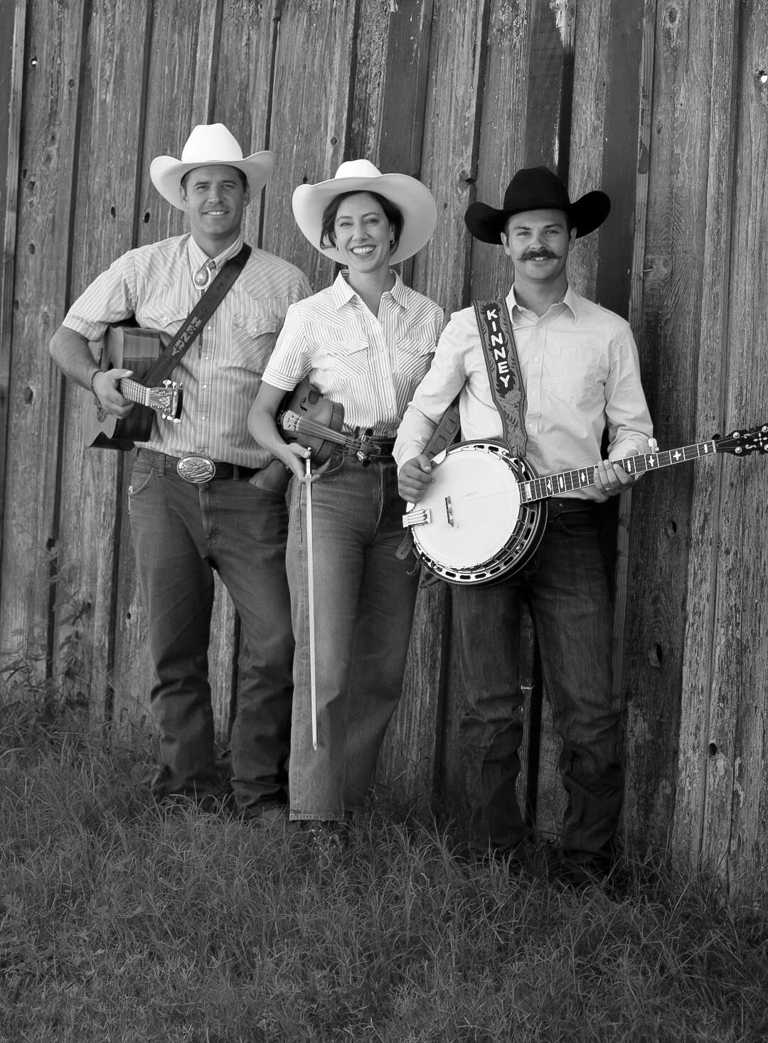 The three members of the Kings River Boys dressed in country western attire standing against a wooden fence, holding  a guitar, a fiddle, and a banjo, smiling at the camera.