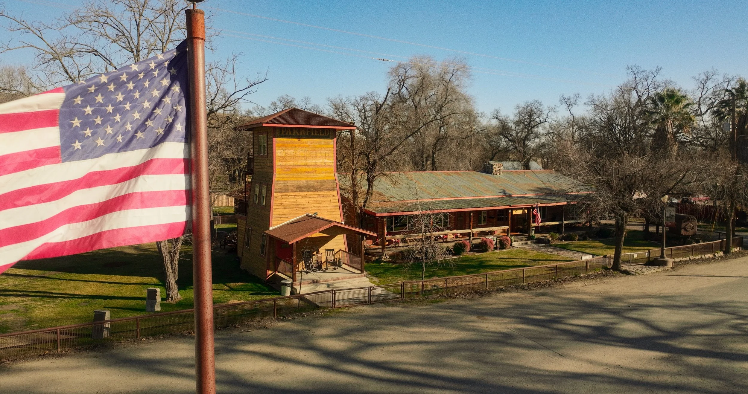 A view of Parkfield Coalinga Road, in Parkfield California. An american flag waving in front of the Parkfield Water Tower with "PARKFIELD" painted on it and trees and the Parkfield Lodge in the background.