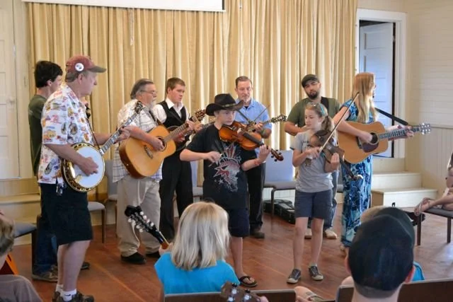 A group of children and a man playing musical instruments performing for an audience in the historic Parkfield Community Hall during the Parkfield Bluegrass Festival