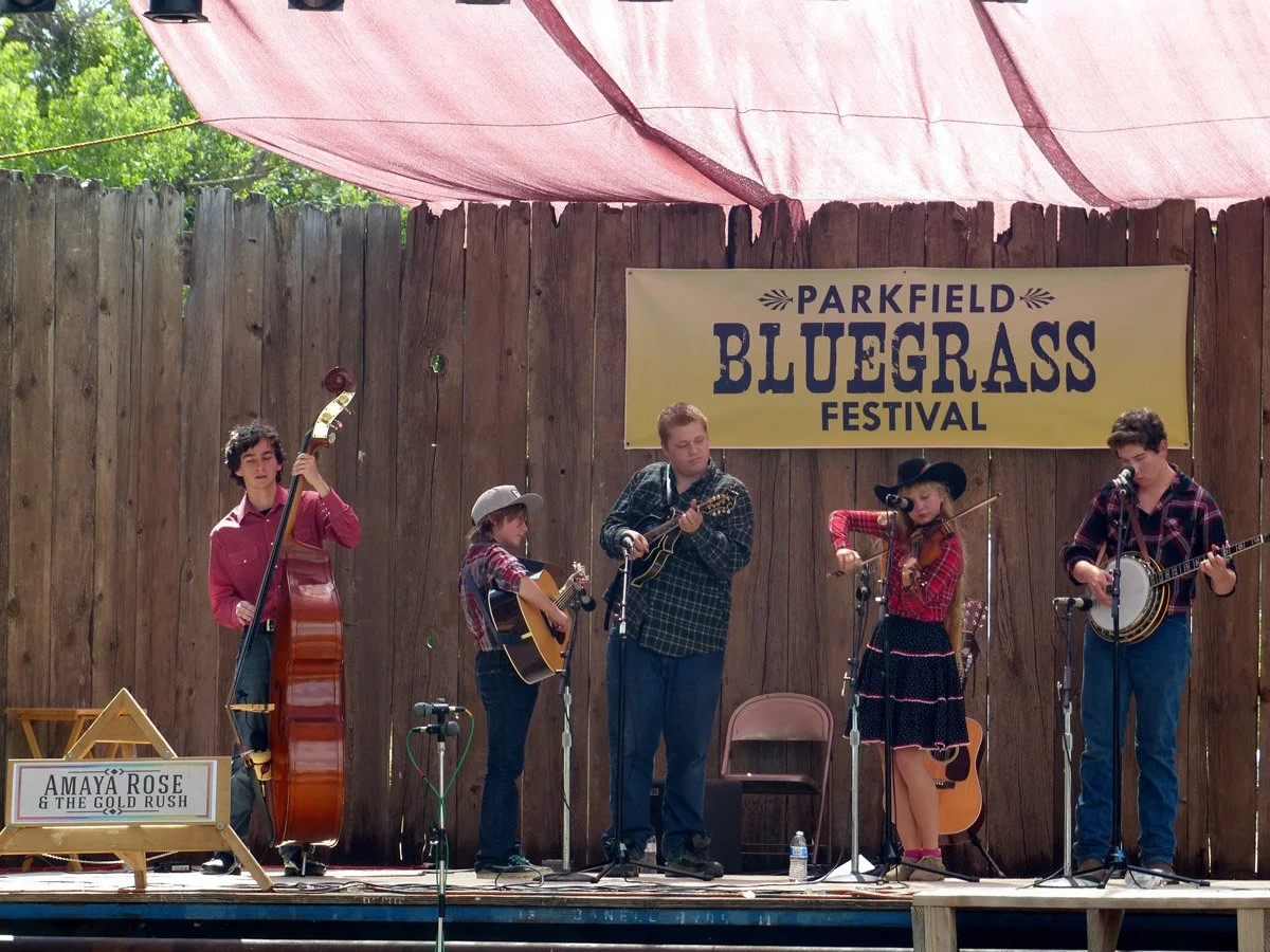 A group of five young musicians performing on a wooden stage at the Parkfield Bluegrass Festival.