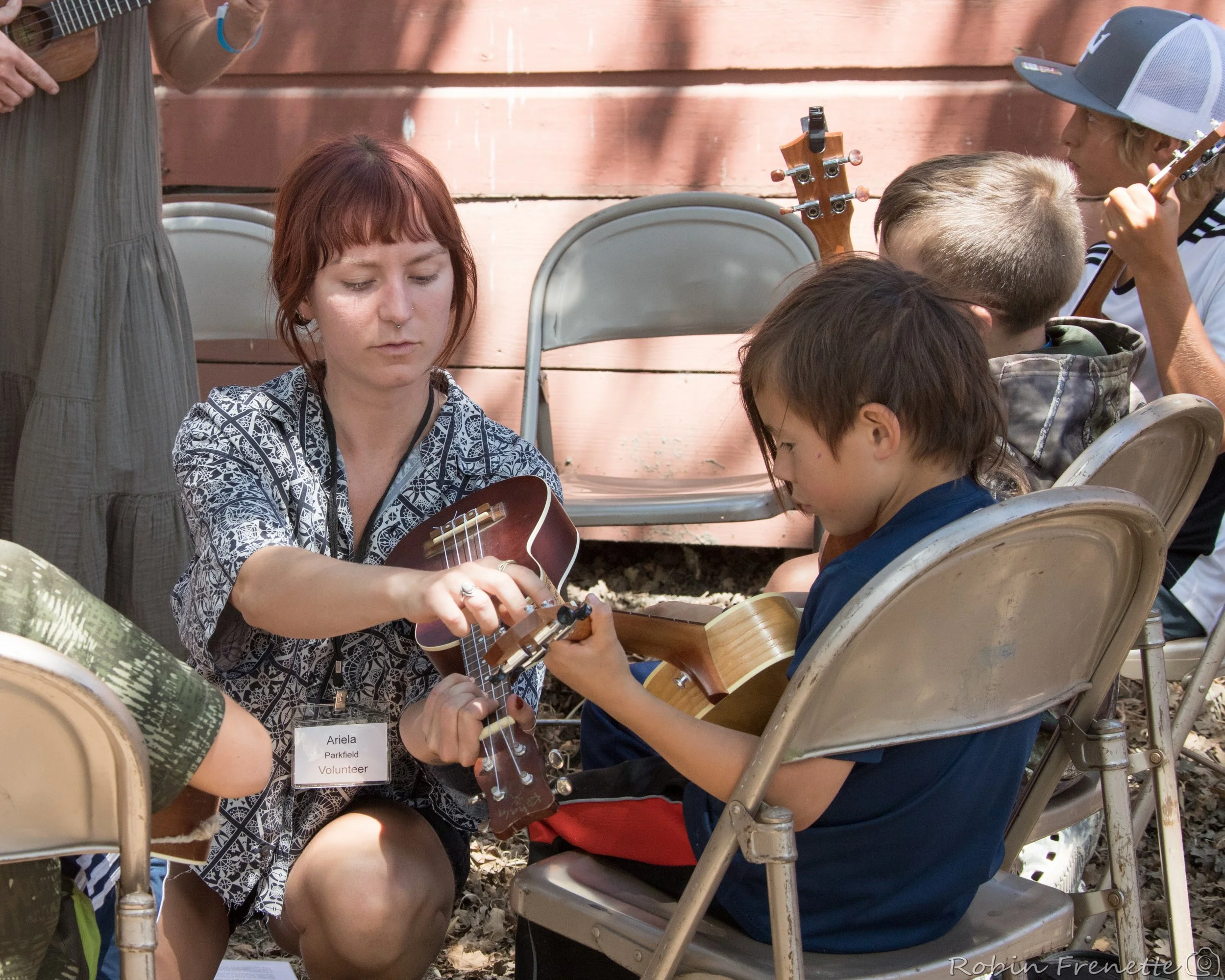 A woman labeled as a volunteer helping children playing string instruments, and singing, at the kids on bluegrass workshop at the Parkfield Bluegrass Festival