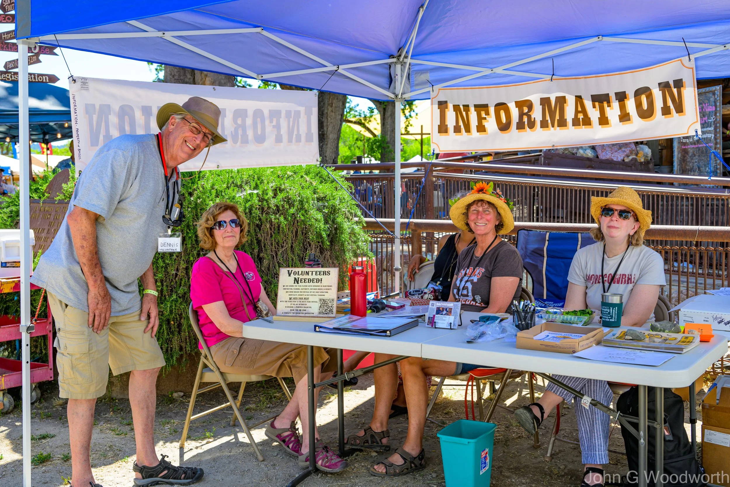 Volunteers standing and smiling at the information booth at the Parkfield Bluegrass Festival, outdoors on a sunny day.