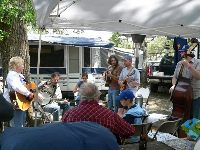 A group of people playing acoustic guitars and a banjo outdoors under a canopy, during the Parkfield Bluegrass Festival