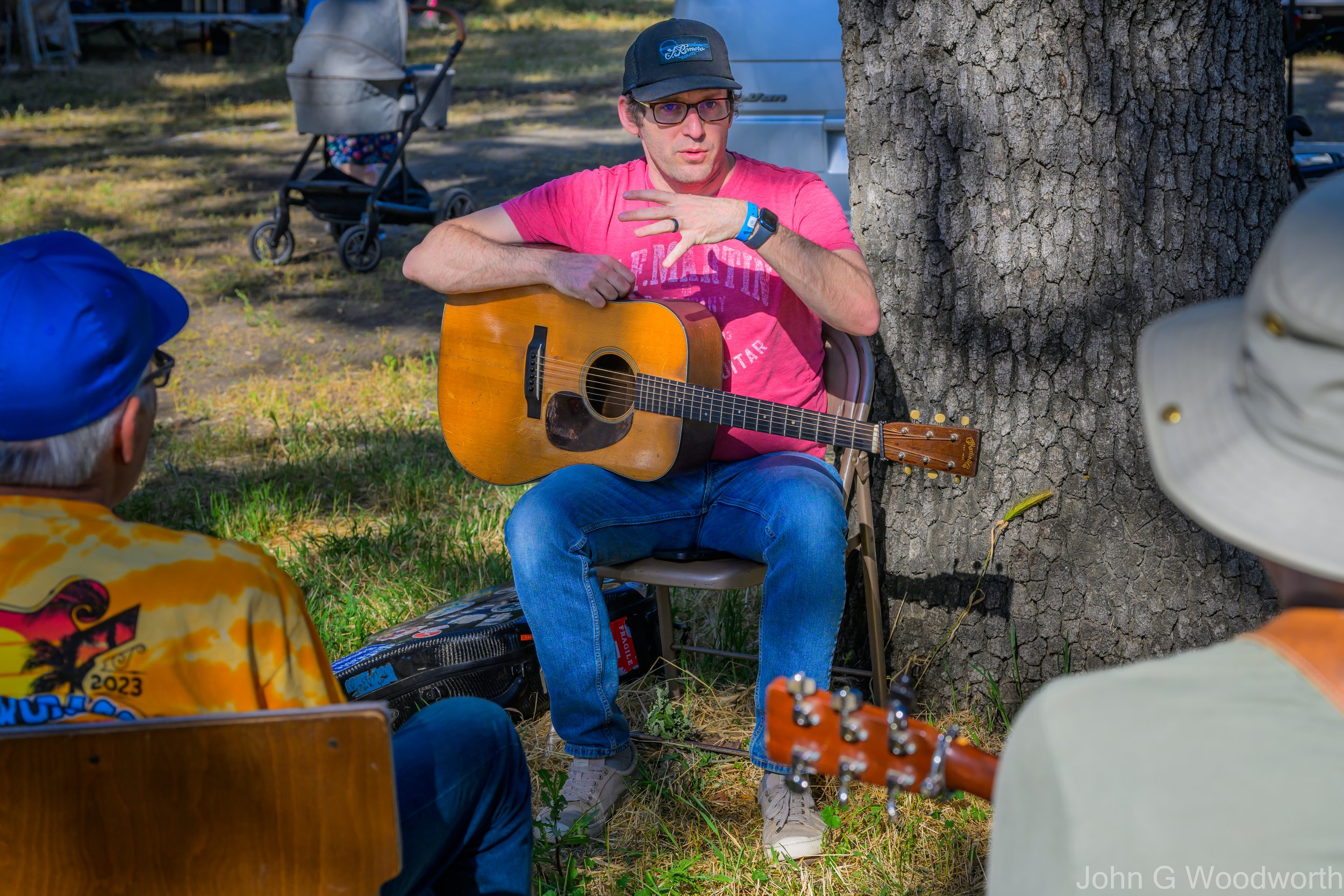 A man sits outdoors under a tree playing an acoustic guitar, with two other men and a woman nearby at a Parkfield Bluegrass Festival workshop. The scene is set in a grassy area with a stroller and a white van in the background.