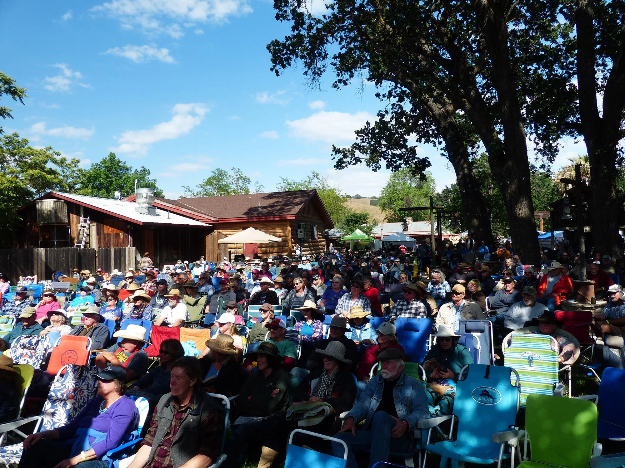 Audience at the Parkfield Bluegrass Festival seated outdoors on lawn chairs under trees, with the Parkfield Cafe, tents and booths in the background.