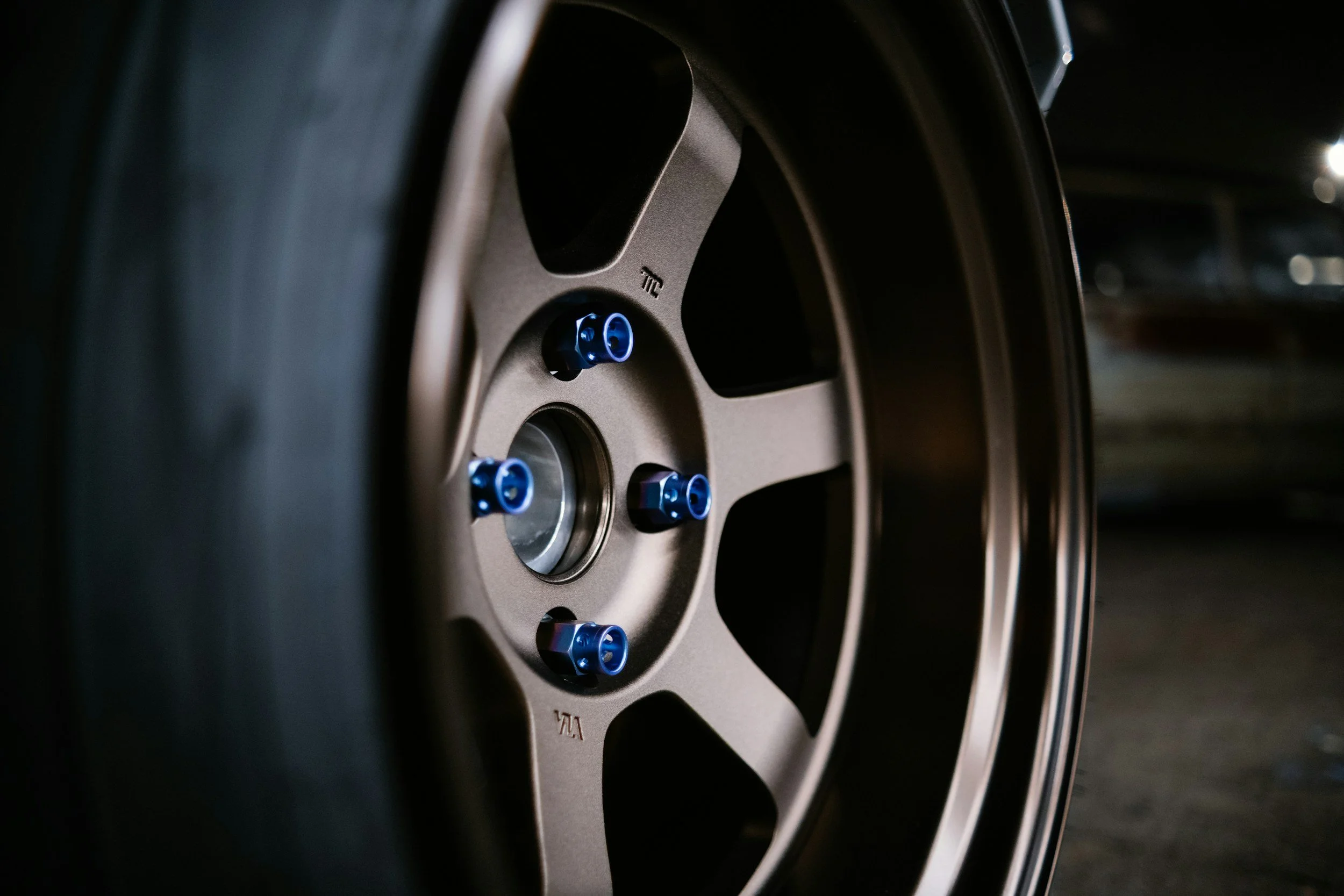 Close-up of a car wheel with a beige rim and blue lug nuts, in a dimly lit environment.