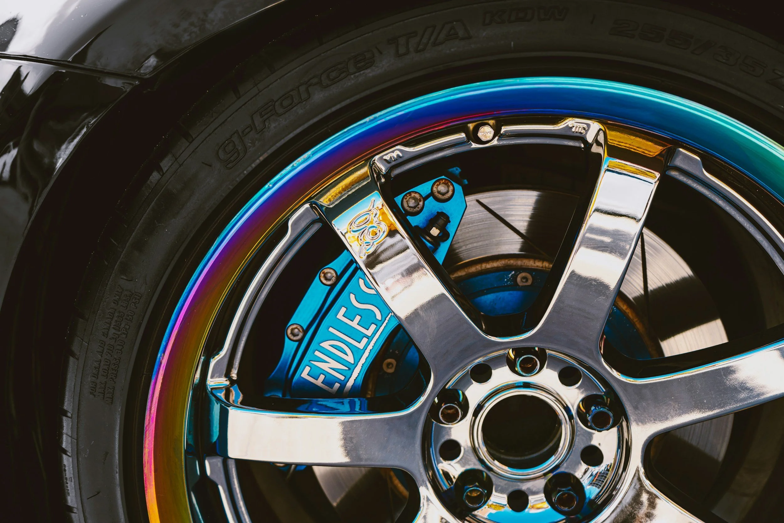Close-up of a racing car wheel showing the rim, tire, and brake components. The rim is shiny chrome, and the brake caliper is painted blue with the word 'ENDLESS' on it. The tire has a rainbow-colored reflective strip along the edge.