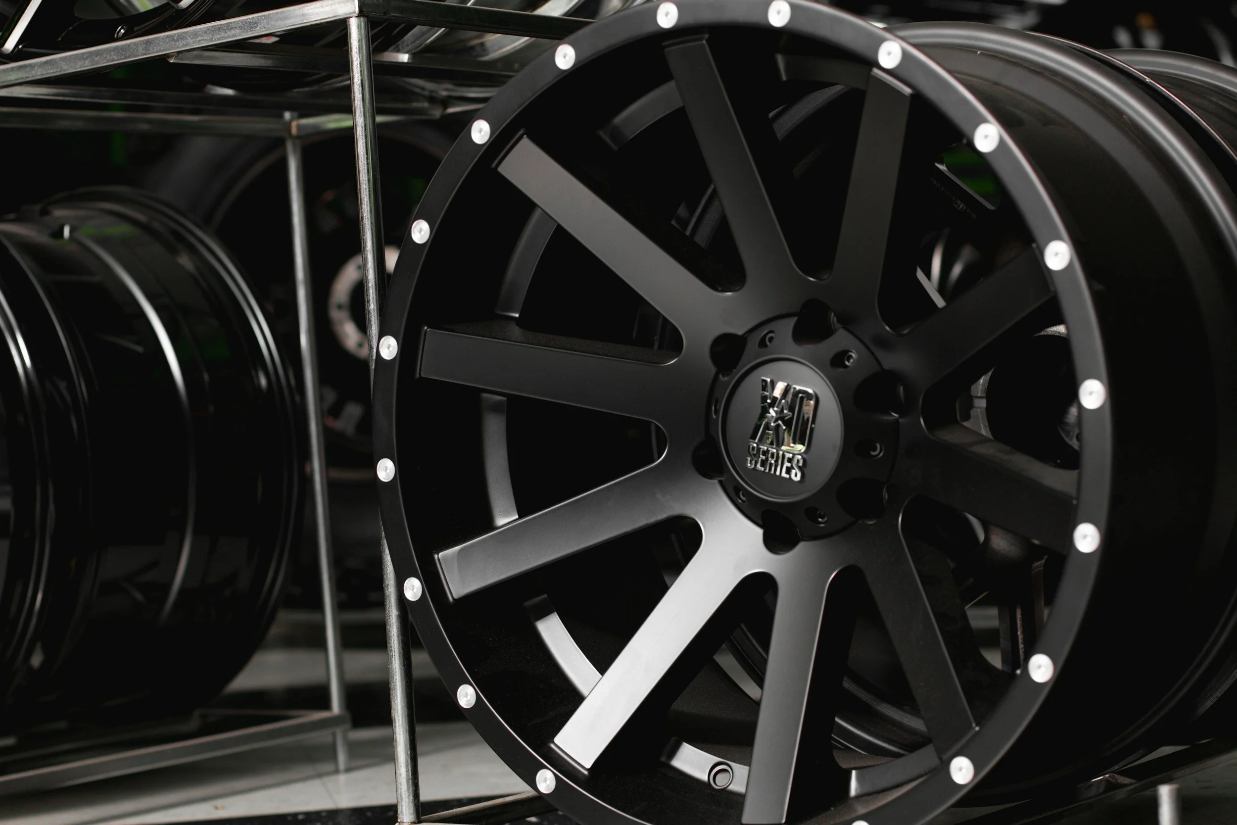 A close-up of black alloy wheels displayed on a metal rack, with a logo in the center reading "K-Series".