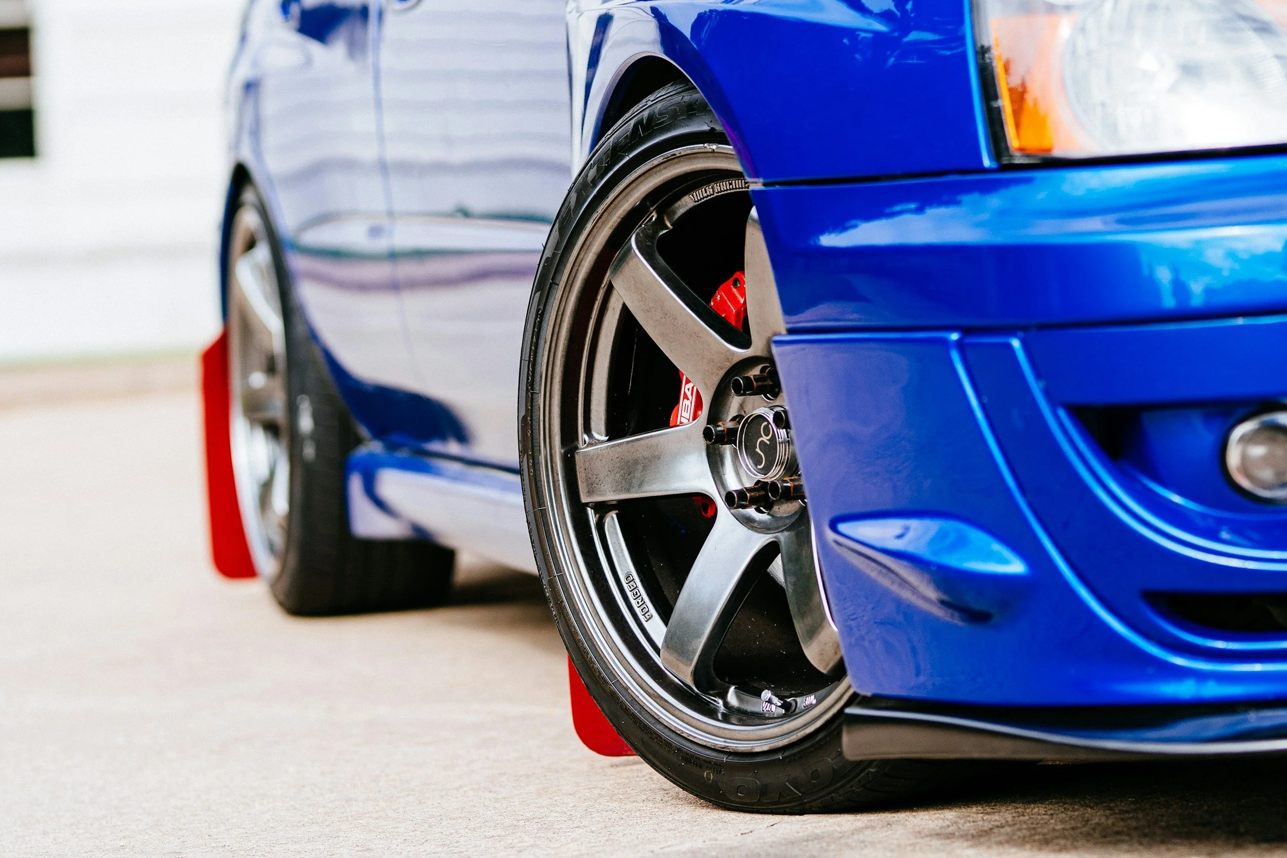 Close-up of a blue sports car's front wheel with a large brake caliper, racing slick tires, and aftermarket rim, parked on concrete.