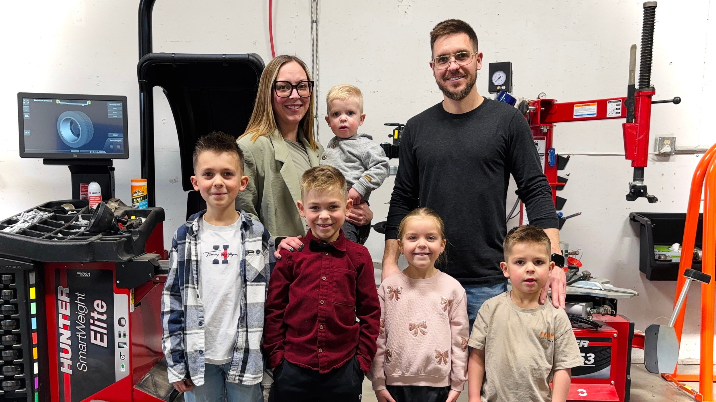 A group of seven people, two adults and five children, standing in a workshop surrounded by tire equipment and tools, smiling at the camera.