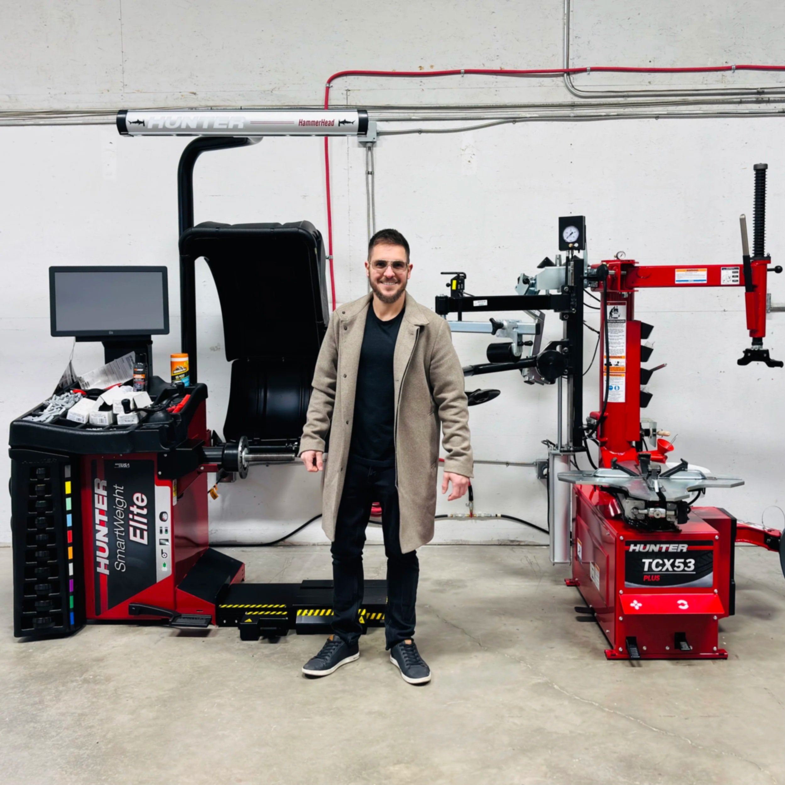 A man wearing a beige coat and black pants standing in front of a wheel balancing machine inside a tire shop in Okotoks, Alberta.