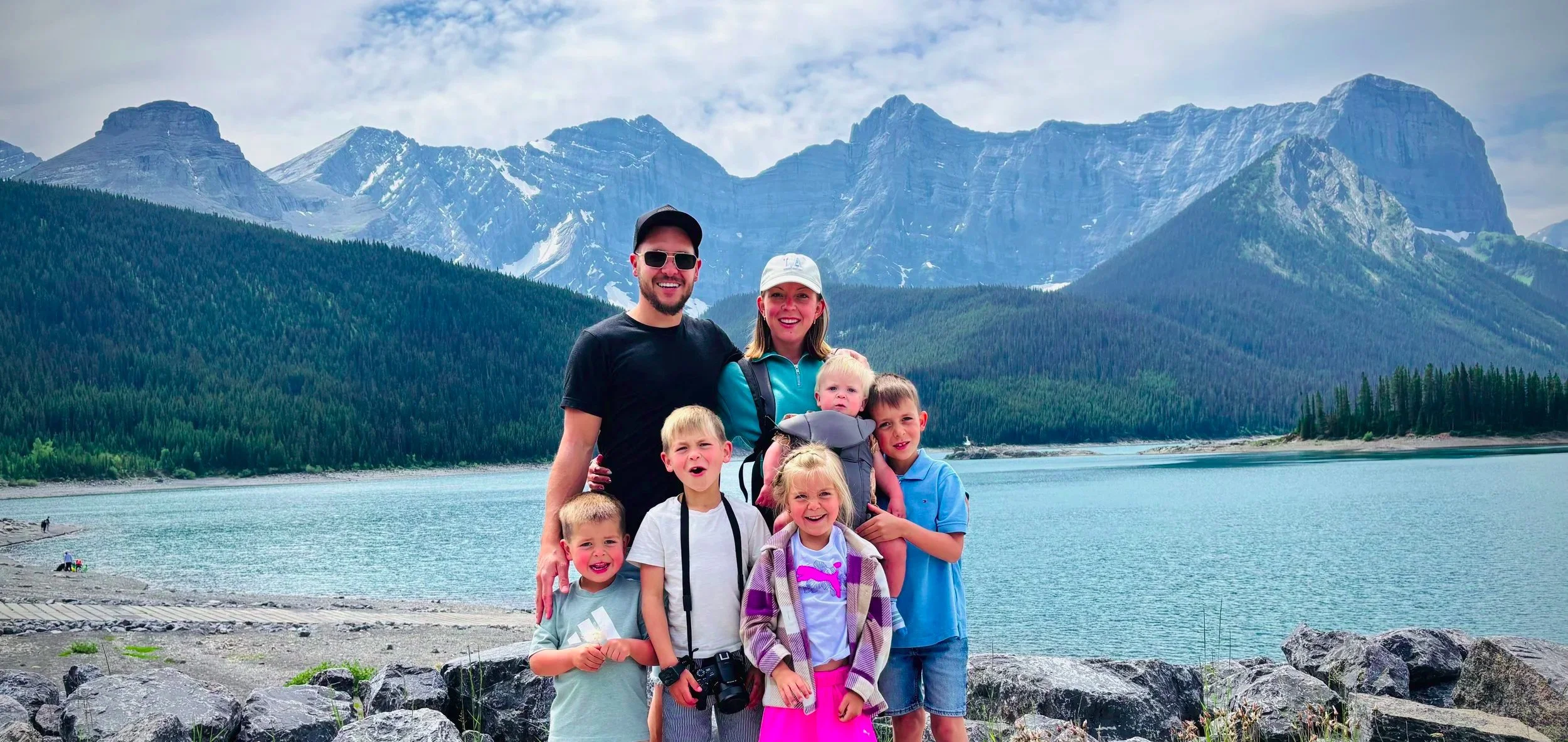 Family of seven, including two adults and five children, standing by a lake with mountains and trees in the background on a cloudy day.