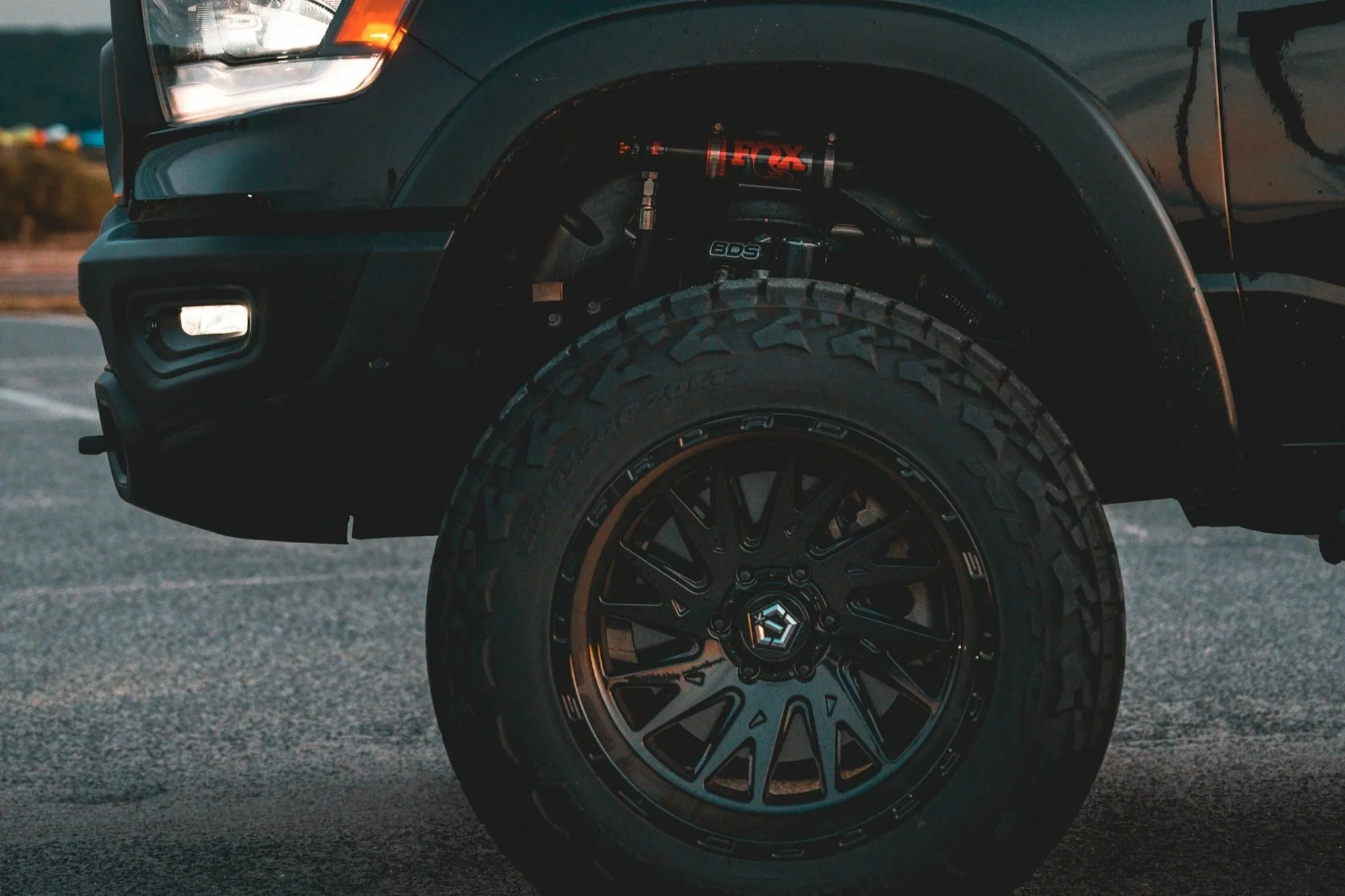 Close-up of a black pickup truck's front wheel and suspension system, including a red FX shock absorber and black custom rim, parked on asphalt.
