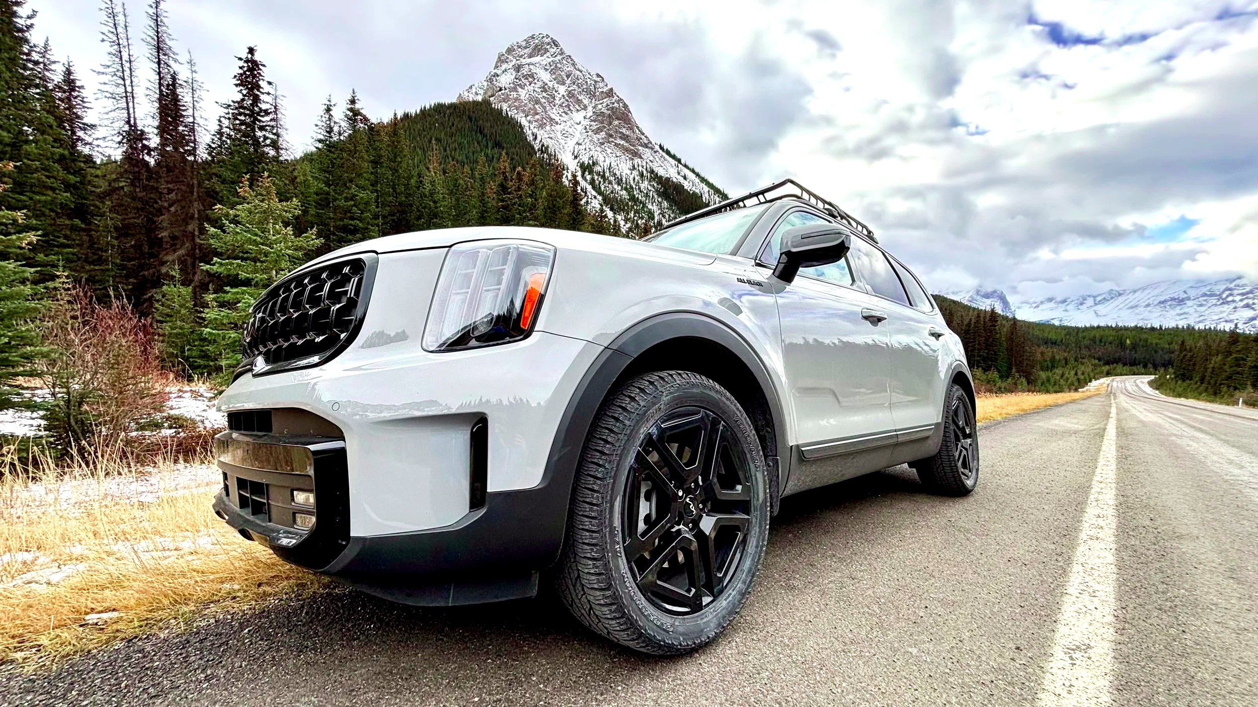 A LIGHT GREY SUV parked on the side of a mountain road with snow-capped peaks and pine trees in the background