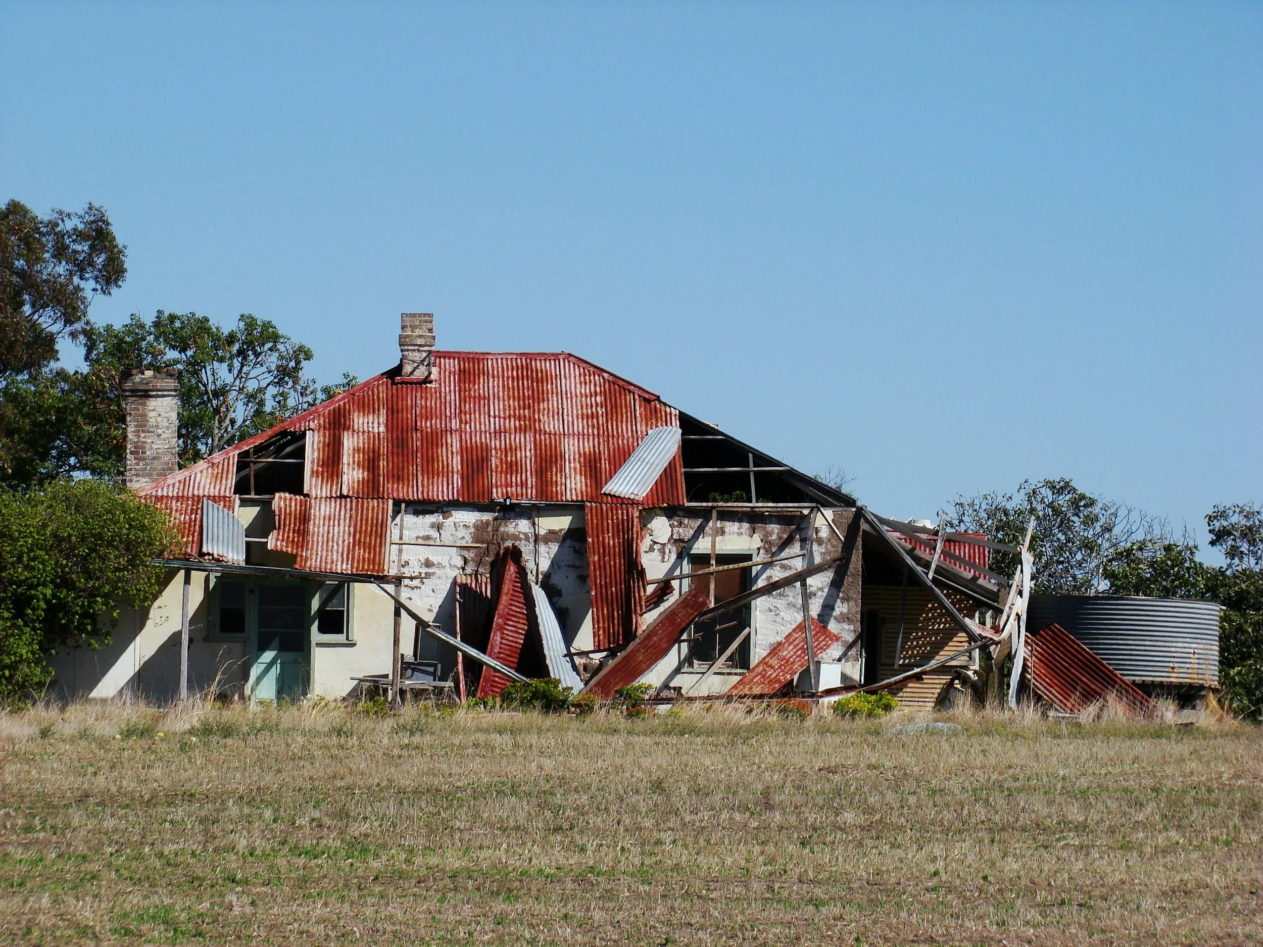 a dilapidated house with a partially collapsed, rusted metal roof, surrounded by sparse grass and trees in the background