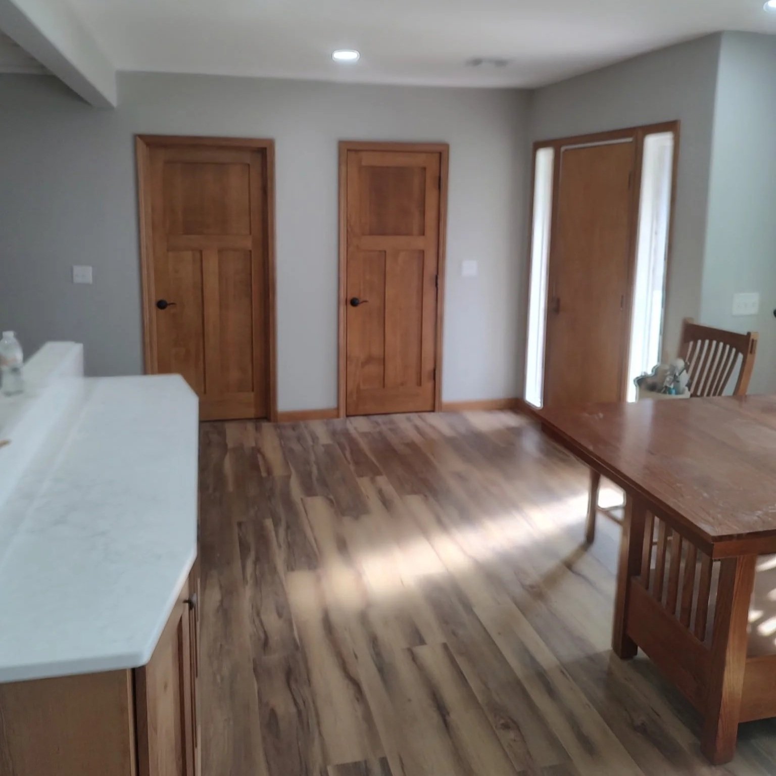 A room with wooden floors, three wooden doors, a wooden dining table, and a chair, with sunlight coming through a door or window on the right side.