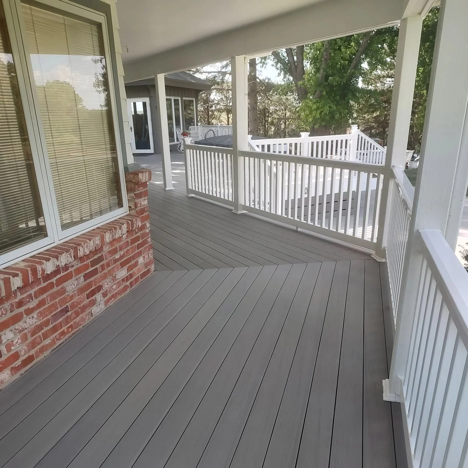 A spacious, covered balcony with gray wooden flooring and white railing, attached to a house with brick and siding exterior, overlooking trees and neighboring houses.