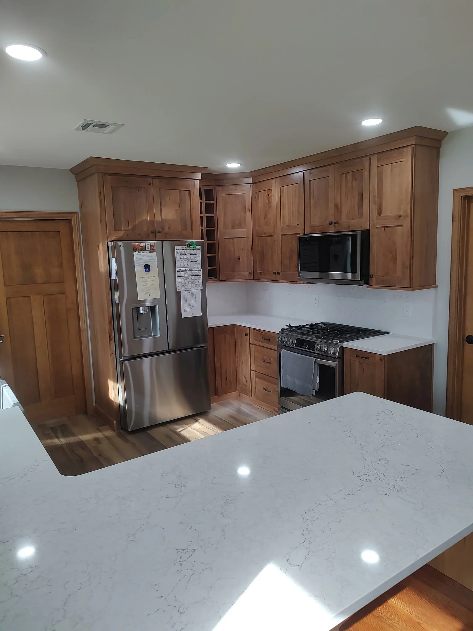 Modern kitchen with wooden cabinets, stainless steel refrigerator, microwave, and oven, white marble countertop, and recessed lighting.