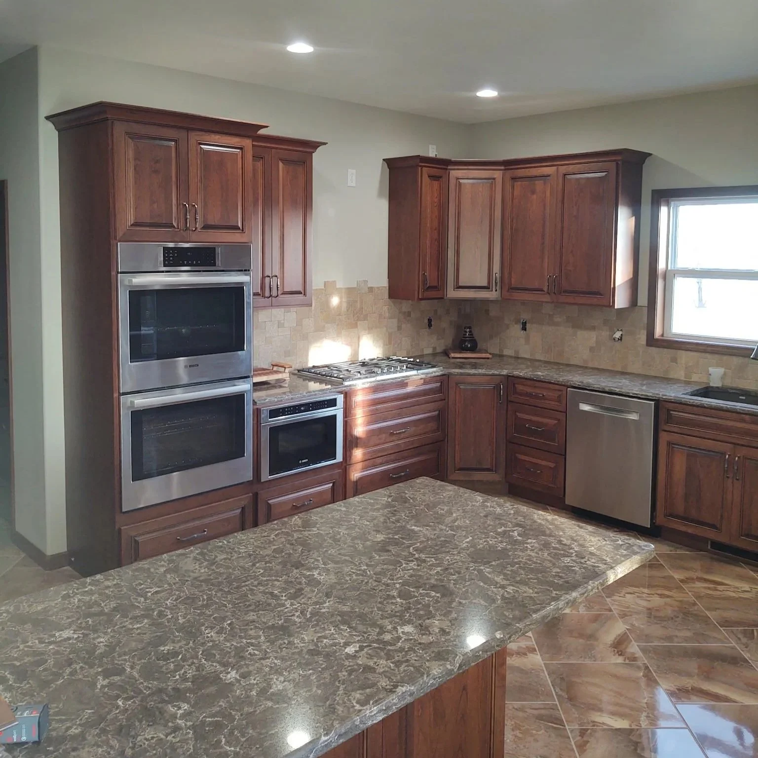 Modern kitchen with wooden cabinets, granite countertops, and stainless steel appliances, including an oven, microwave, dishwasher, and a gas stove, with a window above the sink.