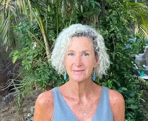 A woman with curly gray hair wearing a sleeveless blue top stands outdoors in front of tropical plants.