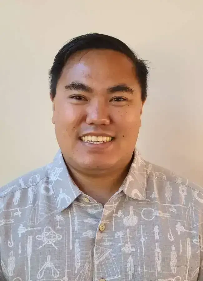 A smiling man with short black hair wearing a light-colored button-up shirt with a pattern of tools and objects, standing against a plain beige wall.