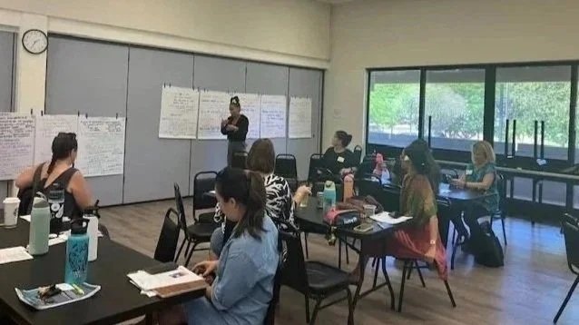 A classroom with women seated at tables, some taking notes, listening to a woman presenting at the front with large papers on the wall behind her. Large windows reveal greenery outside.