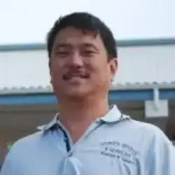 A man with short dark hair smiling outdoors, wearing a light-colored polo shirt, with a building and sky in the background.