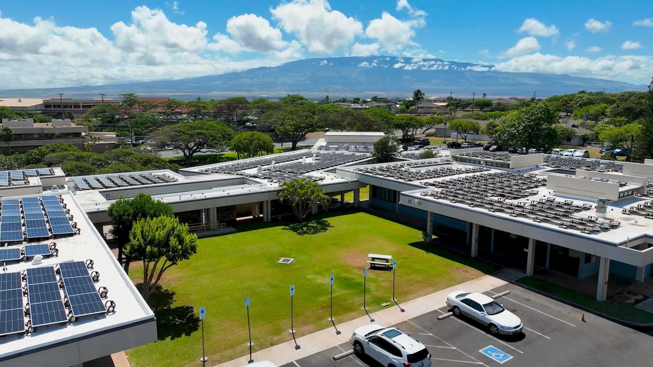 Aerial view of a campus with solar panels on the rooftops, a grassy area with trees, and a parking lot with a few cars, with a mountain and partly cloudy sky in the background.