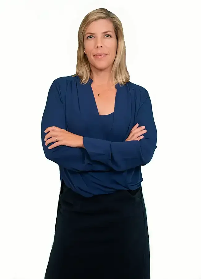 A woman with blonde hair, wearing a blue top and black skirt, standing with her arms crossed against a white background.