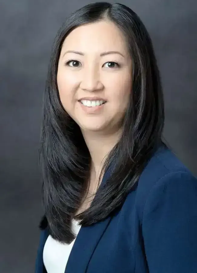 A woman with long black hair, smiling, wearing a navy blazer and white top against a dark background.