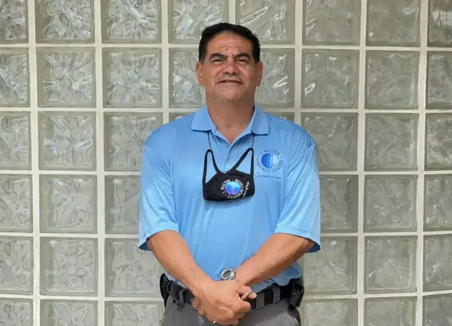A man with dark hair and a mustache smiling, standing in front of a glass block wall. He is wearing a light blue polo shirt with a logo, a face mask hanging around his neck, and a watch on his left wrist.