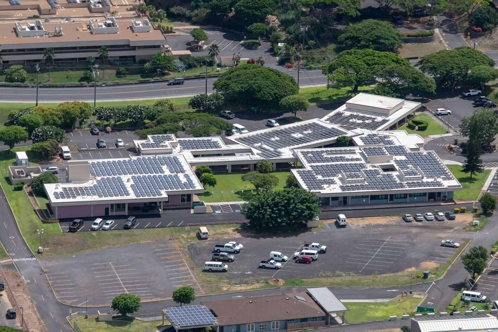 Aerial view of a building complex with multiple rooftops covered in solar panels, surrounded by parking lots, trees, and roads.