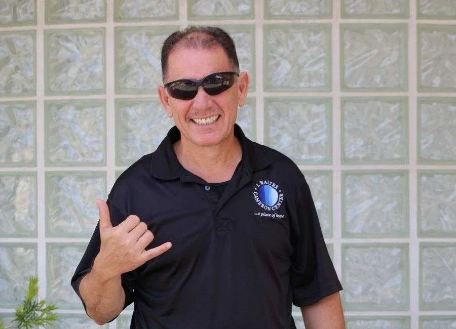 A smiling man with short dark hair, wearing sunglasses and a black polo shirt, making a shaka sign with his right hand, standing in front of a tiled wall.