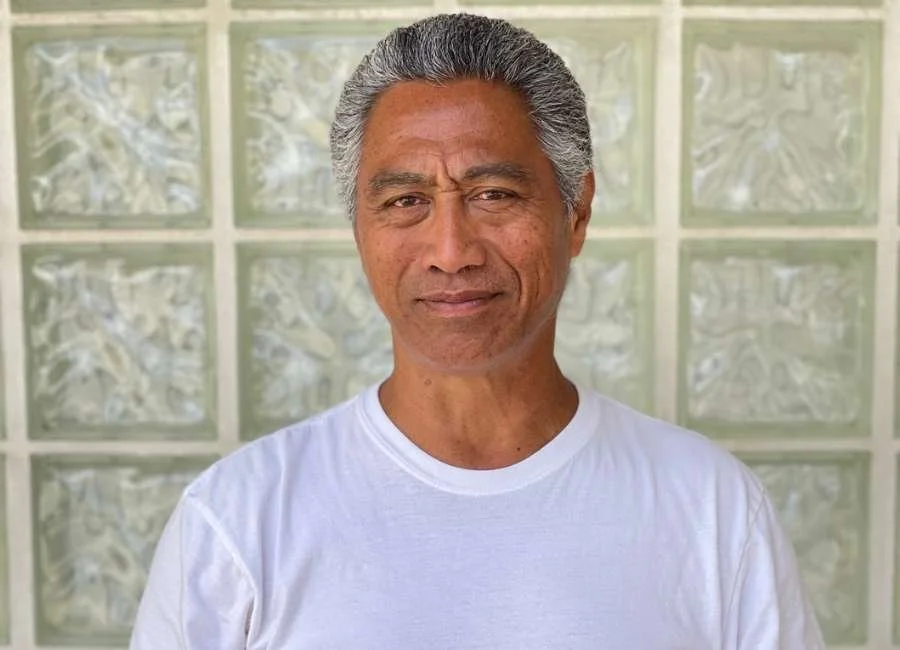 Portrait of an older man with gray hair, wearing a white t-shirt, standing in front of a glass block wall.
