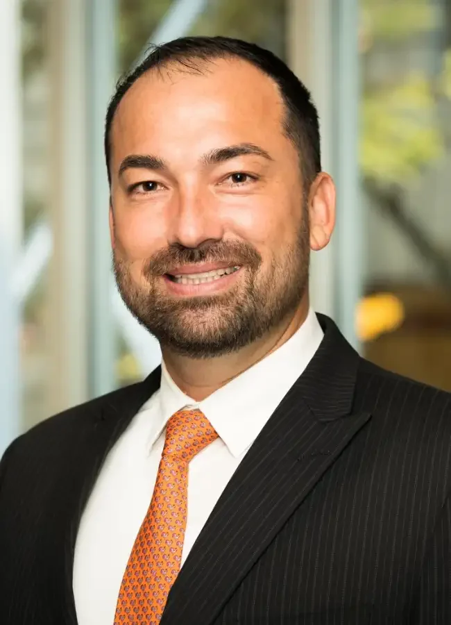 Headshot of a man in a business suit smiling, with short dark hair, a beard, and a patterned orange tie, in an office setting.