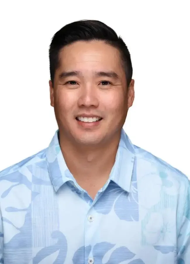 Close-up of a smiling Asian man with short black hair wearing a light blue patterned shirt, against a plain white background.