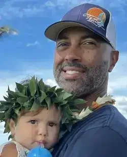 An adult man with a beard wearing a baseball cap and a young girl with a leaf crown on her head, outdoors in a sunny environment.