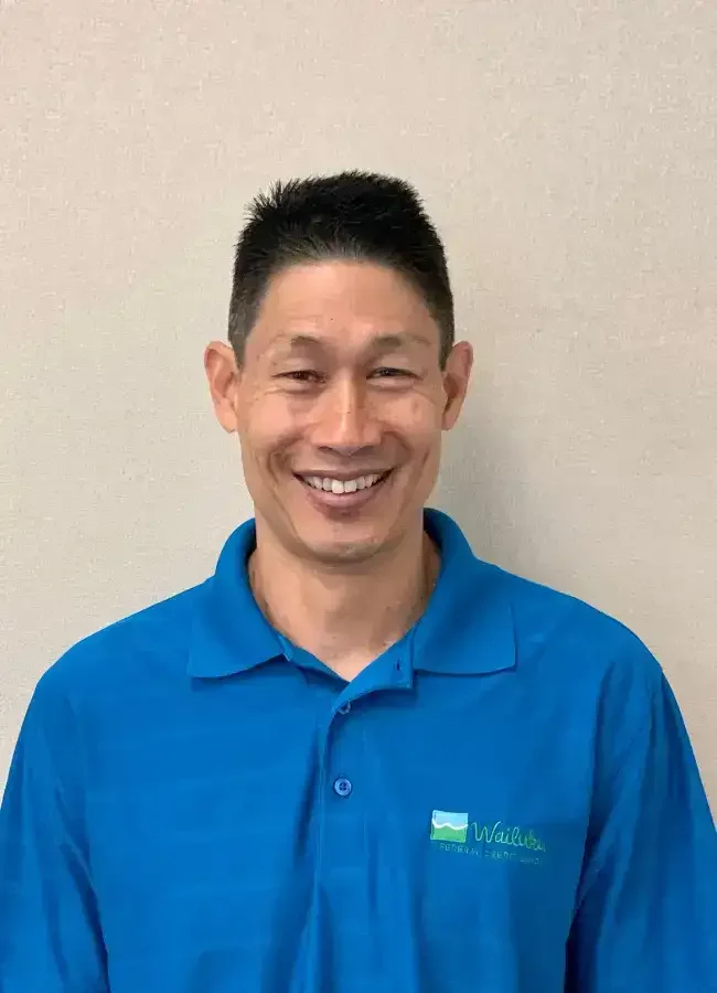 Man wearing a blue polo shirt with a logo, smiling, against a plain wall.