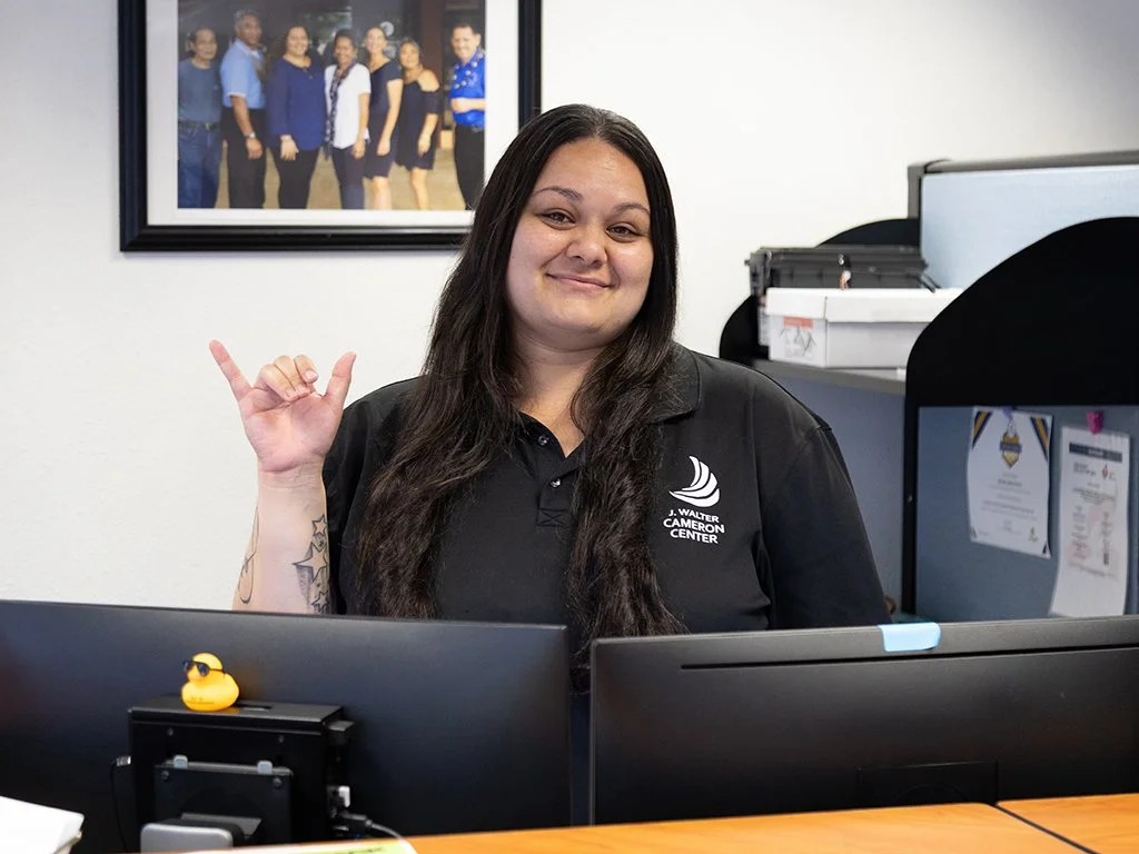 A woman with long dark hair, smiling, making a 'hang loose' gesture with her left hand, sitting at a desk with computer monitors in an office. She is wearing a black polo shirt with the logo 'J. Walter Cameron Center'.