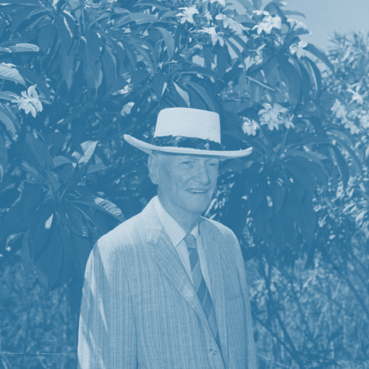JW Cameron wearing a white hat, suit, and tie, standing outdoors with trees and flowers in background.