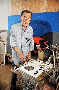 An elderly man with glasses smiling while standing at an art table with paintbrushes, paints, and art supplies in front of a blue backdrop.