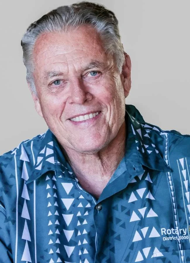 A smiling elderly man with gray hair wearing a blue patterned shirt with a Rotary District logo.