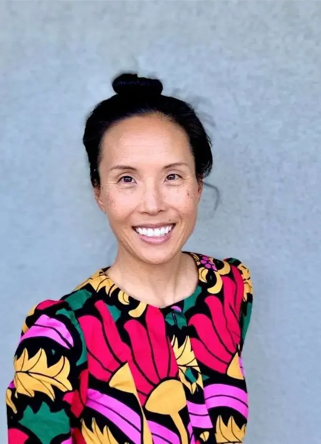 A woman with dark hair styled in a top knot, smiling and wearing a colorful, patterned top, standing against a plain gray wall.