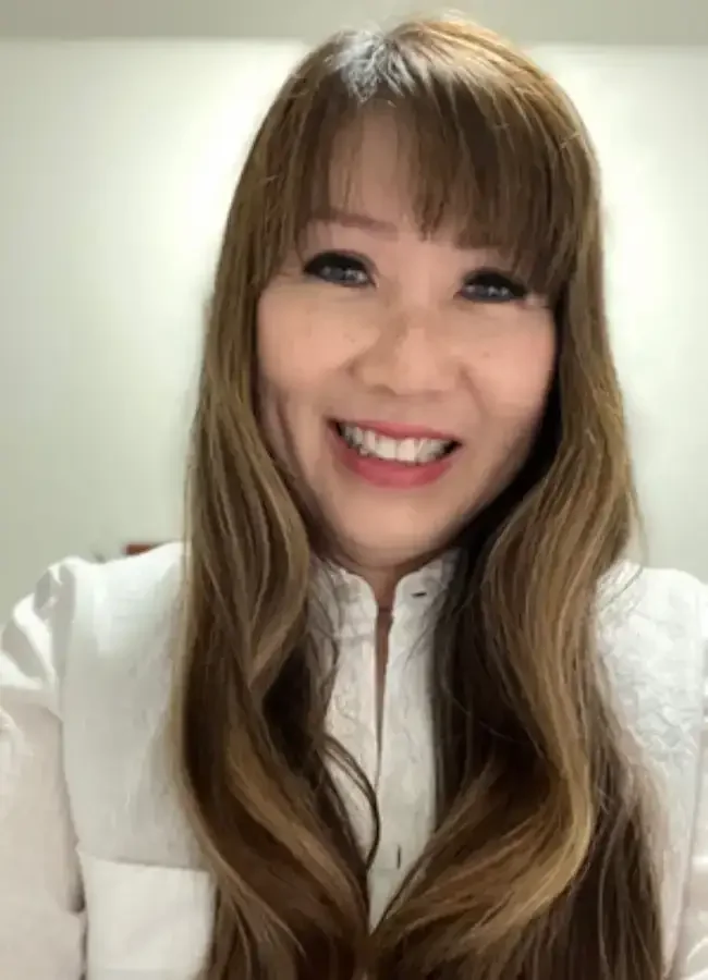 A woman with long, wavy brown hair and bangs, smiling and wearing a white top with a zipper.