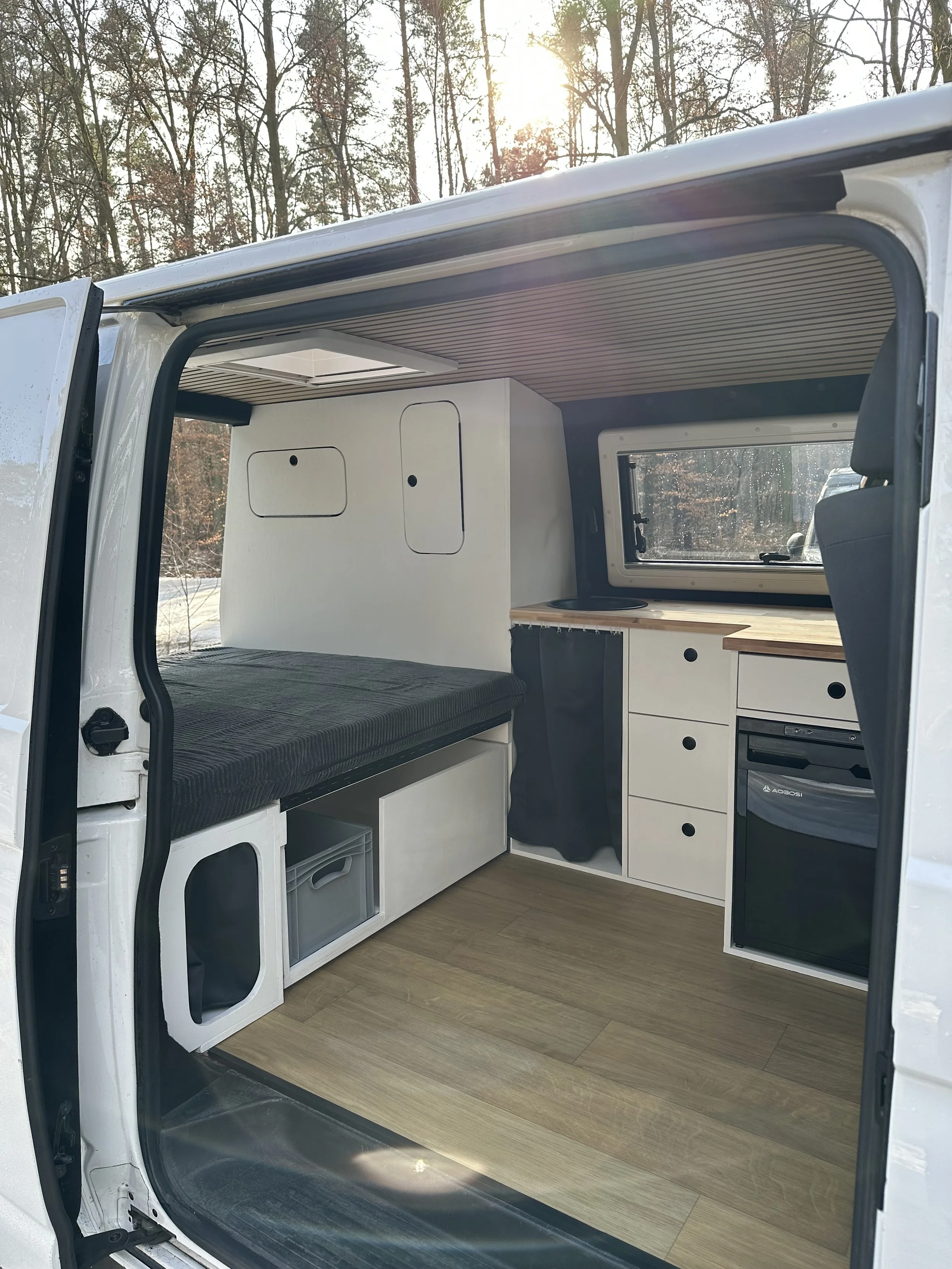 Innenansicht eines umgebauten Fahrzeugs mit Schlafbereich, Kamins, kleinen Stauraumfächern und Küchenbereich, bei Sonnenlicht im Freien