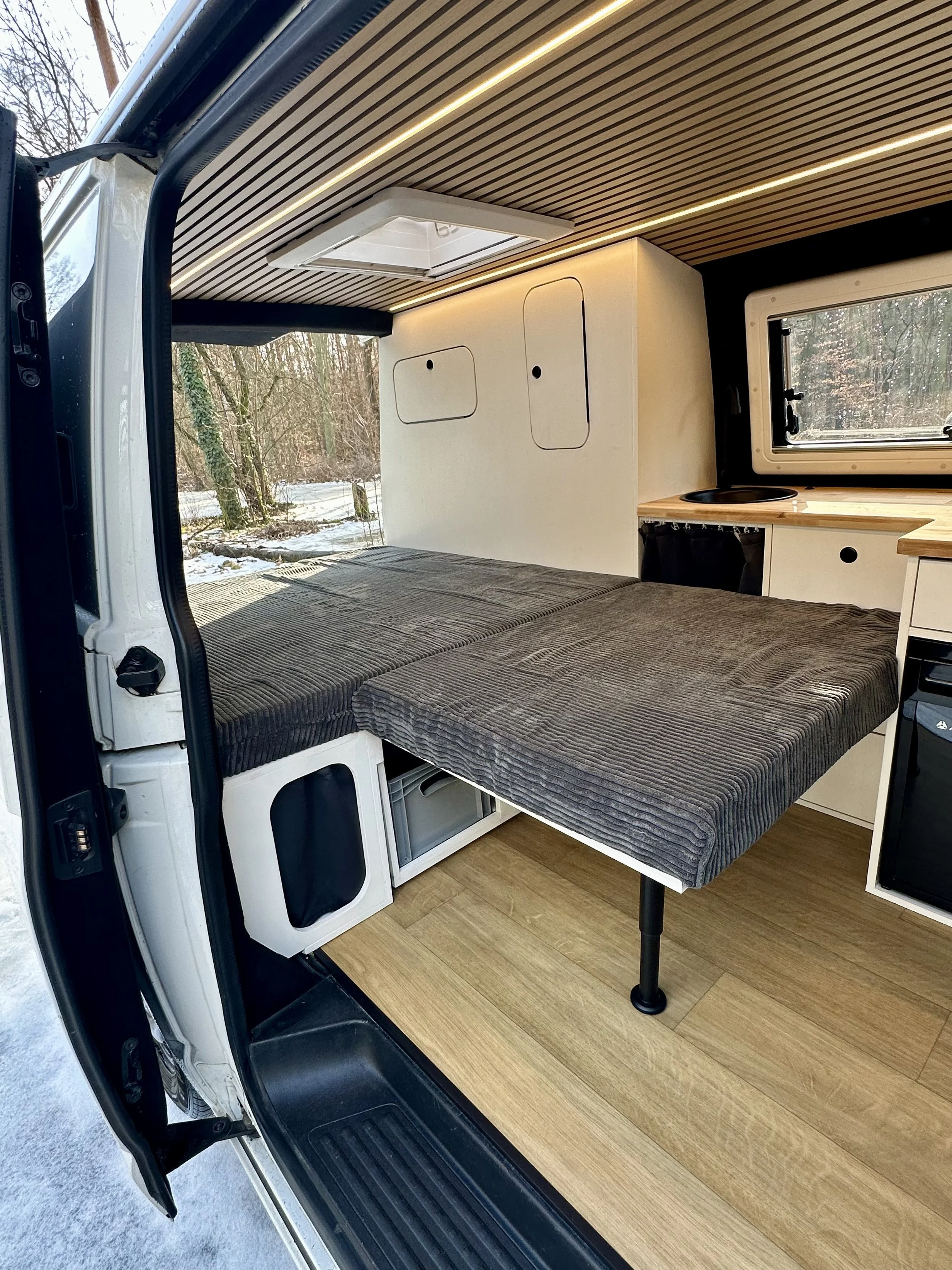 Innenansicht eines Camper Vans mit umgebauter Schlafgelegenheit, Holzdecke, Seitentasche, Fenster und Küchenbereich