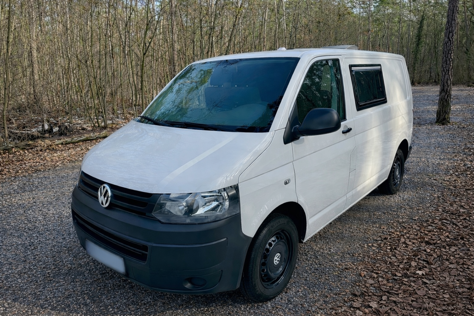 Weiße Volkswagen Transporter-Kastenwagen auf einem Kiesweg in einem Wald mit Bäumen und Laub auf dem Boden.