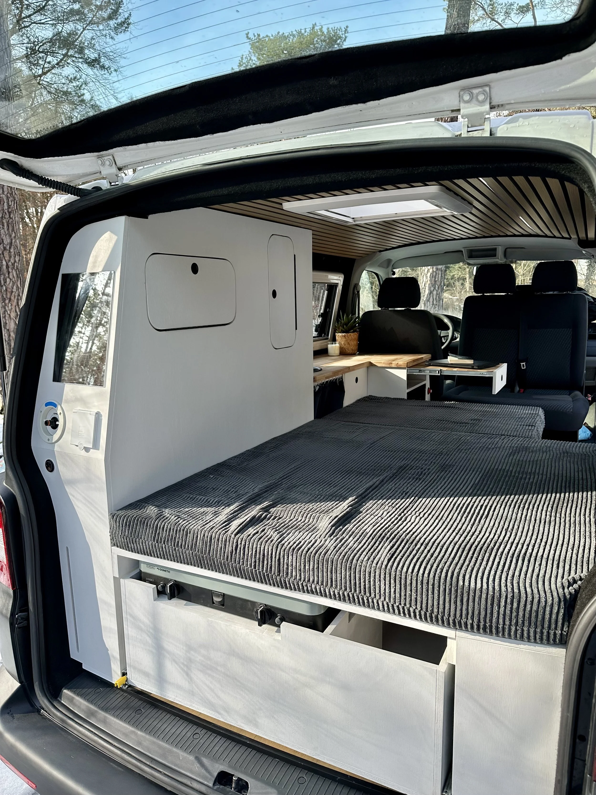 Innenraum eines umgebauten Vans mit Bett, Stauraum und Arbeitsbereich, im Hintergrund die Fahrerkabine, bei Tageslicht