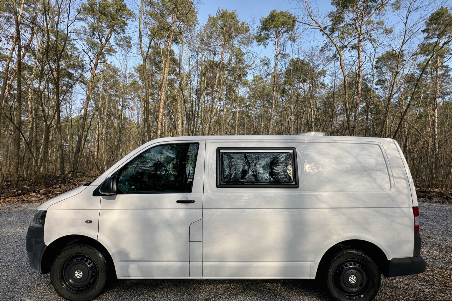 Weiße Lieferwagen vor einem Wald bei Sonnenlicht, mit reflektierenden Fenstern.